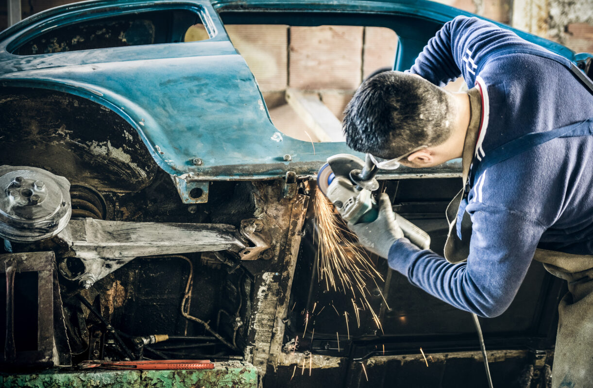 Young man mechanical worker repairing old vintage car body with electric grider in messy garage - Work safety with protection wear - Niche expertise concept with guy working with technical equipment